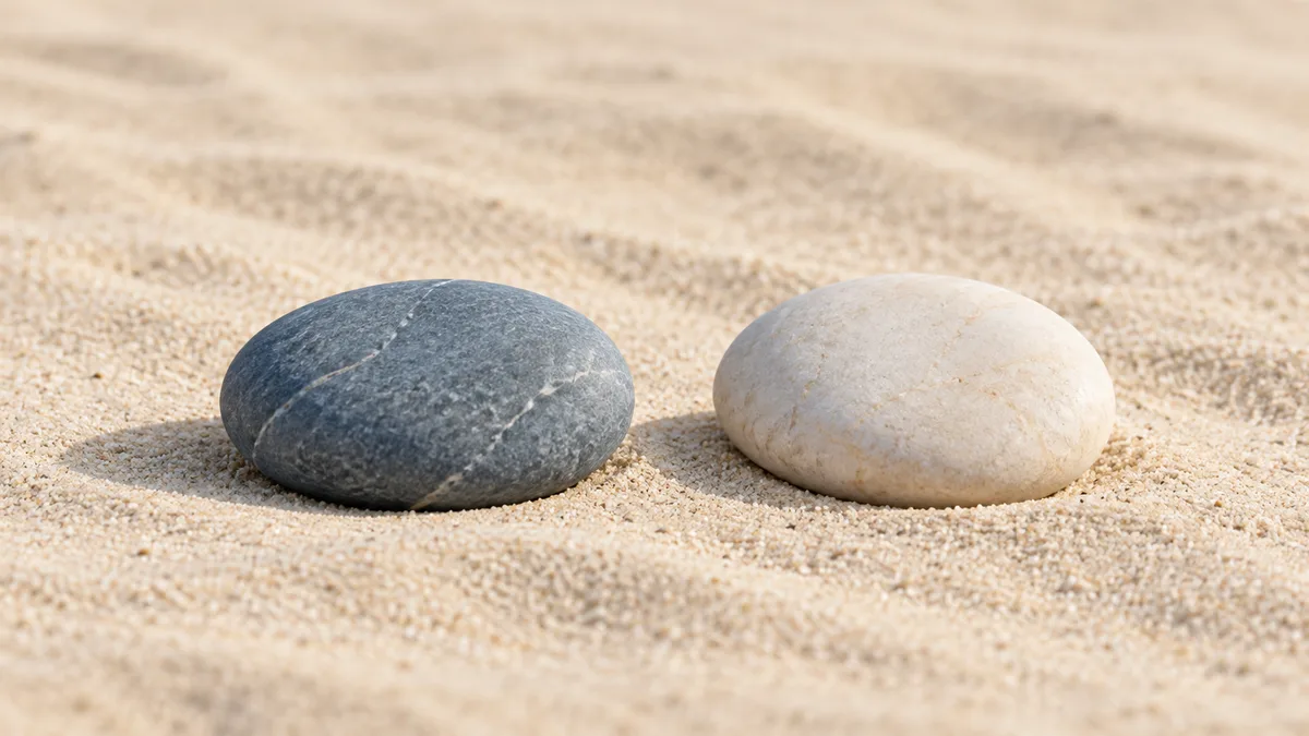 Two smooth river stones, one blue-grey and one ivory, side by side on pale sand.